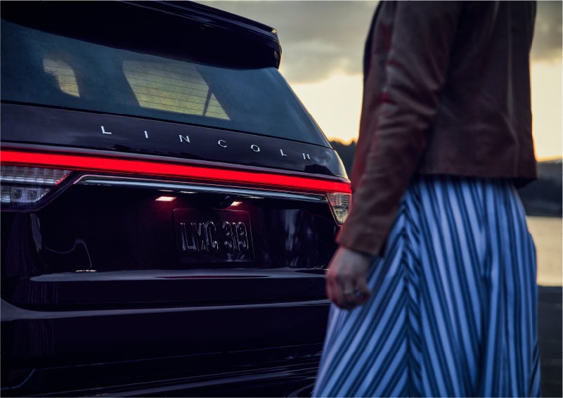 A person is shown near the rear of a 2023 Lincoln Aviator® SUV as the Lincoln Embrace illuminates the rear lights | Thomasville Lincoln in Thomasville GA