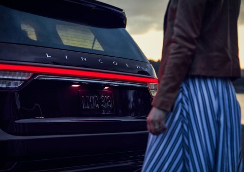 A person is shown near the rear of a 2024 Lincoln Aviator® SUV as the Lincoln Embrace illuminates the rear lights | Thomasville Lincoln in Thomasville GA