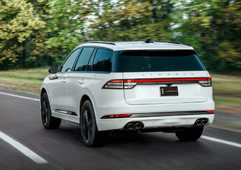 A white 2026 Lincoln Aviator® SUV is being driven away down a road lined with colorful trees. | Thomasville Lincoln in Thomasville GA